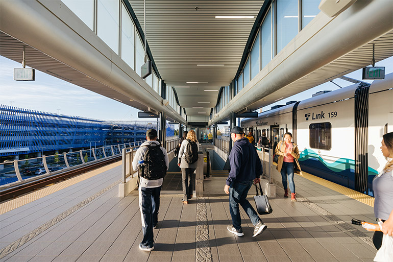 People walking on a light rail platform towards two side-by-side escalators. A light rail train waits in the background.